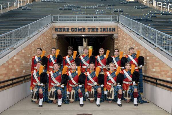 Thirteen members of the Irish Guard, in full red and blue tartan uniform, pose beneath the 